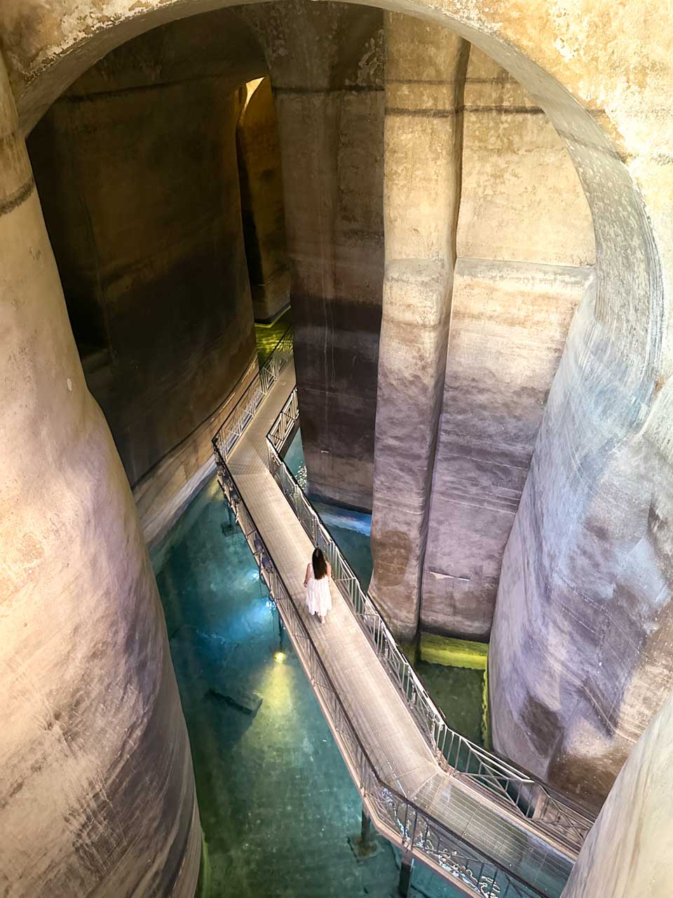 A woman in a white maxi dress walking across the suspended walkway inside the Palombaro Lungo cistern in Matera