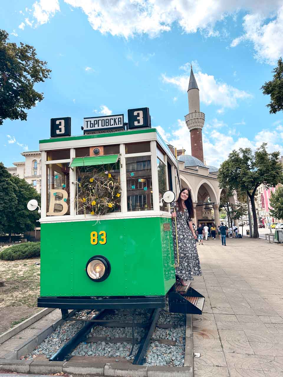 Woman posing on the steps of a vintage green tram with the Banya Bashi Mosque behind her