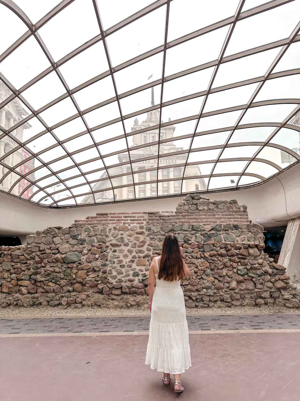 Woman looking at the ancient Serdica ruins beneath the glass dome, with the former Communist Party House visible above