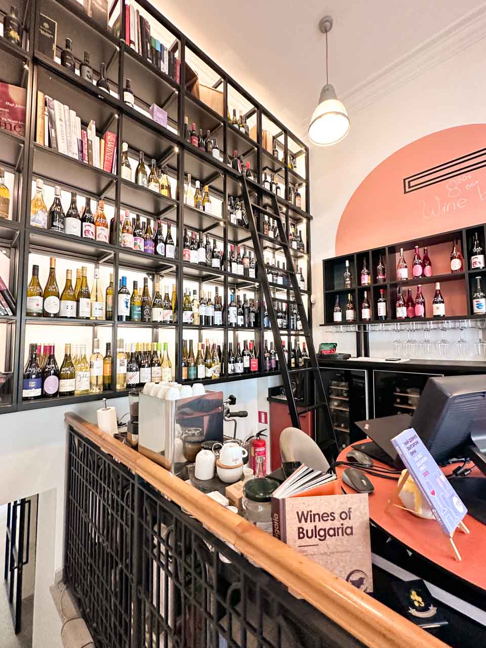 Floor-to-ceiling shelves filled with Bulgarian wine bottles inside Wine Generator in Sofia with a rolling ladder and counter in front