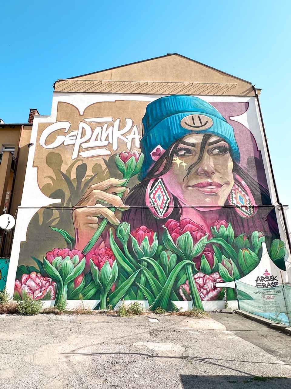 A colourful mural in Sofia showing a woman in a blue beanie holding a tulip, surrounded by pink flowers