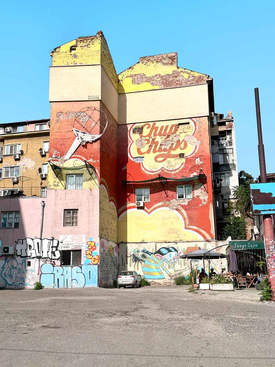 A weathered red and yellow building covered in murals and graffiti in Sofia, with outdoor seating beside it