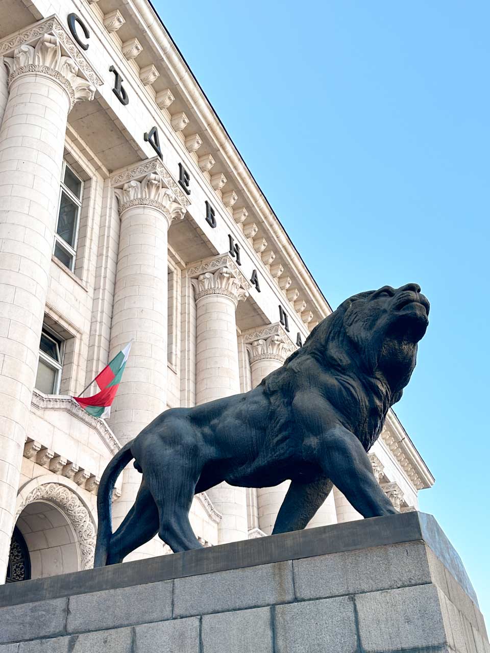 Bronze lion statue outside the Palace of Justice in Sofia, with tall stone columns and a Bulgarian flag behind