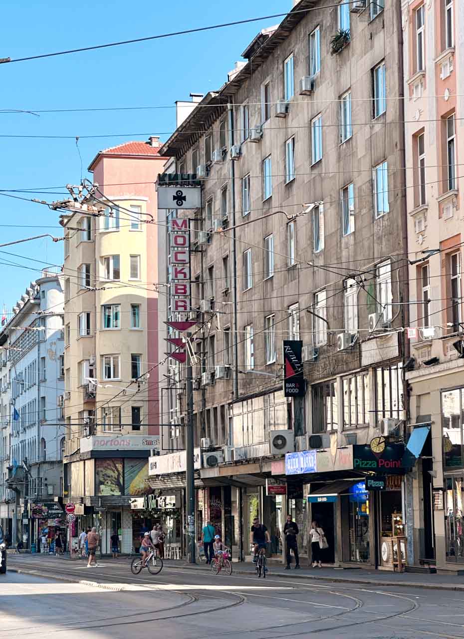 A busy street in central Sofia lined with shops and residential buildings, with tram tracks below and wires overhead