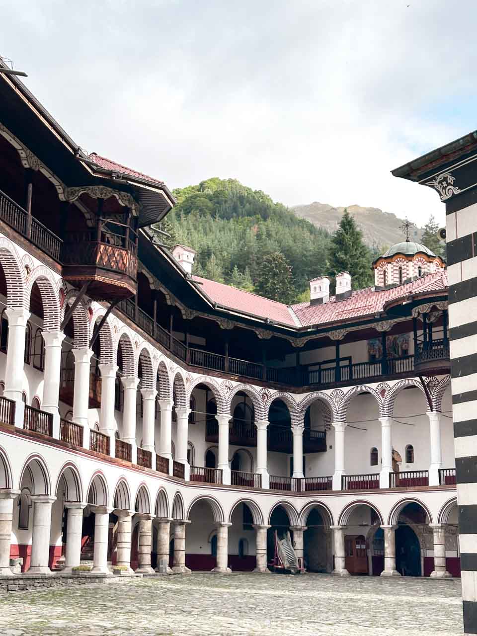 Courtyard of Rila Monastery with rows of striped arches and wooden balconies, and the Rila Mountains in the background