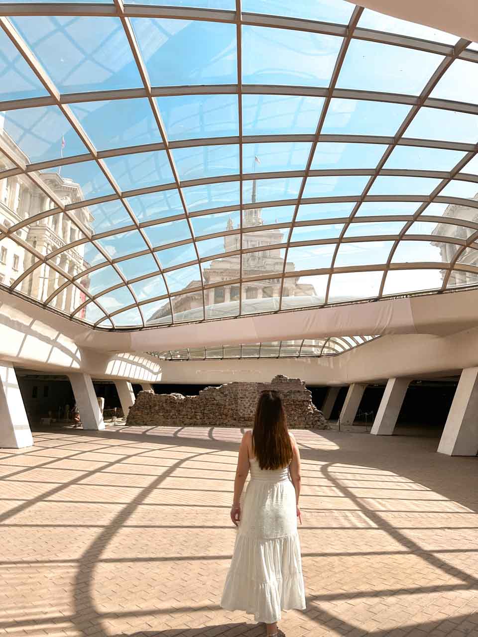 A woman in a white maxi dress walking towards the ancient Serdica ruins beneath a glass dome in central Sofia