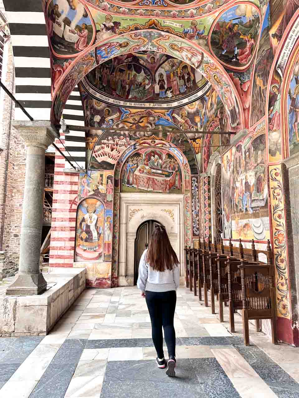 Woman walking beneath colourful religious frescoes outside the church at Rila Monastery, Bulgaria