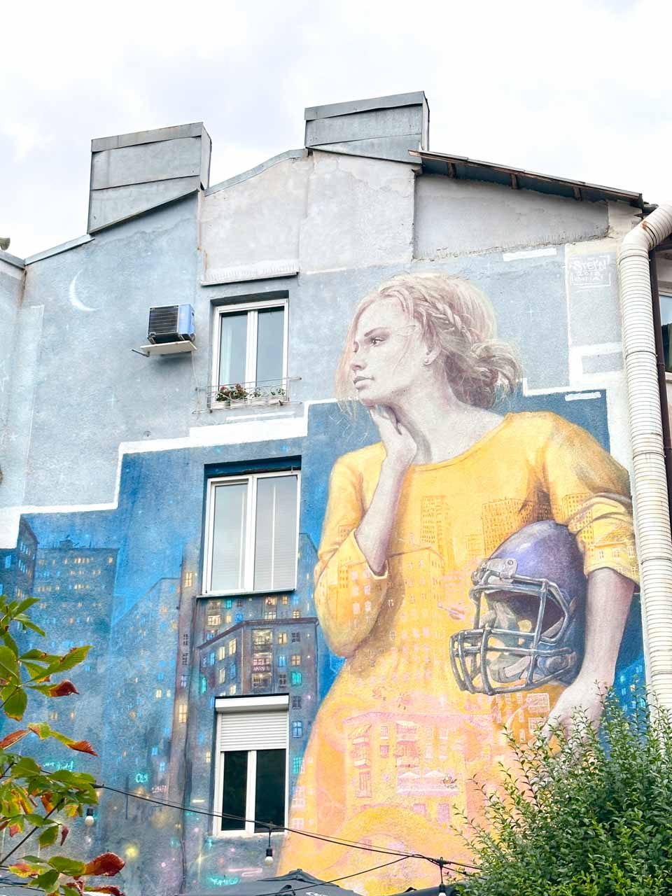 Large street art mural of a woman holding an American football helmet on the side of a building in Sofia