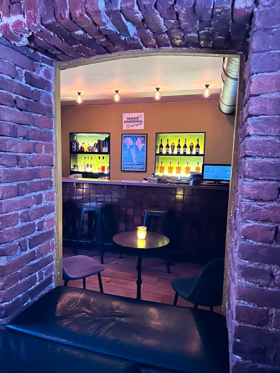 The cosy interior of 5L Speakeasy Bar in Sofia, seen through a brick archway with the bar and illuminated bottle shelves in the background