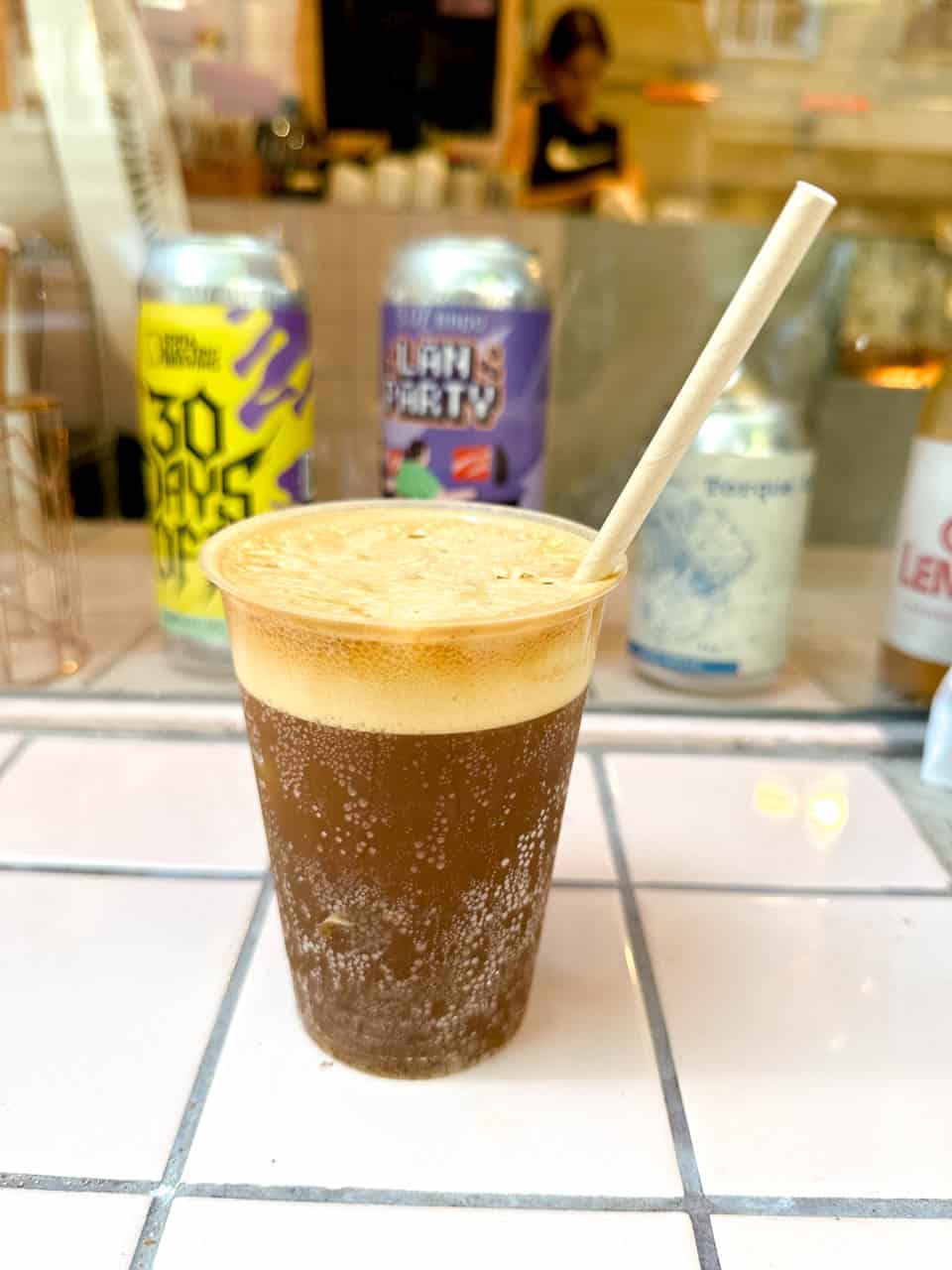 Cold brew coffee served in a plastic cup, with colourful cans lined up behind it at &hlyab (&bread) in Sofia